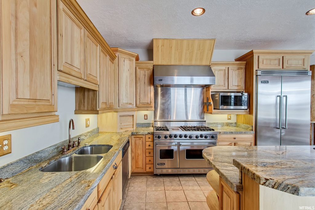 Kitchen featuring wall chimney exhaust hood, light stone countertops, built in appliances, and sink