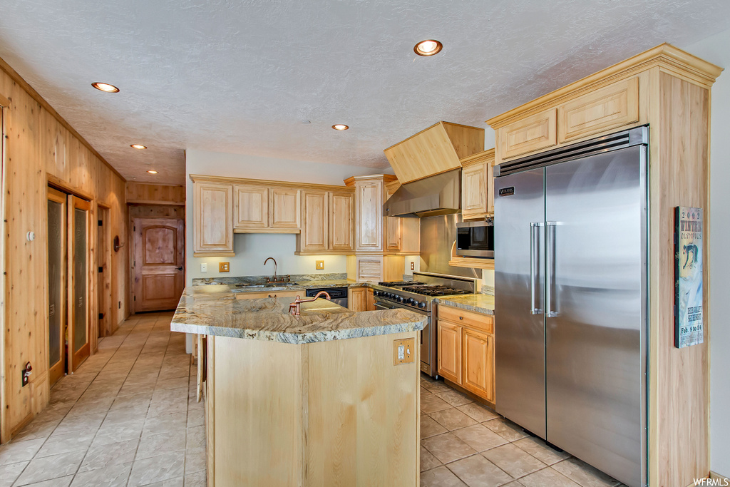 Kitchen featuring premium appliances, sink, light stone countertops, wooden walls, and light brown cabinetry