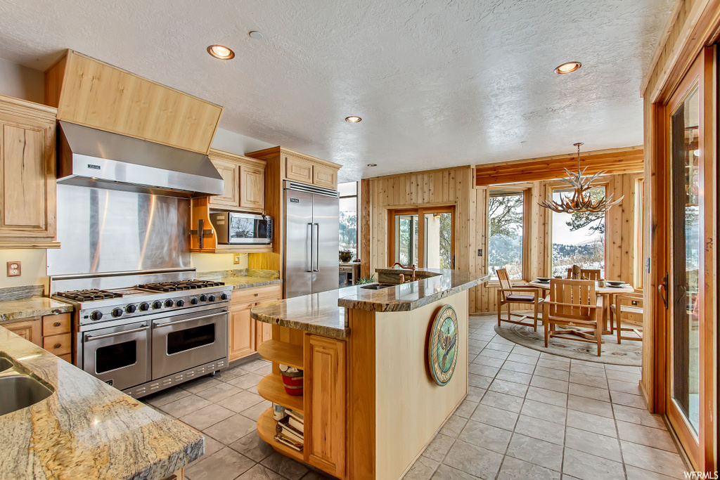 Kitchen featuring light stone counters, wall chimney exhaust hood, built in appliances, and a kitchen island