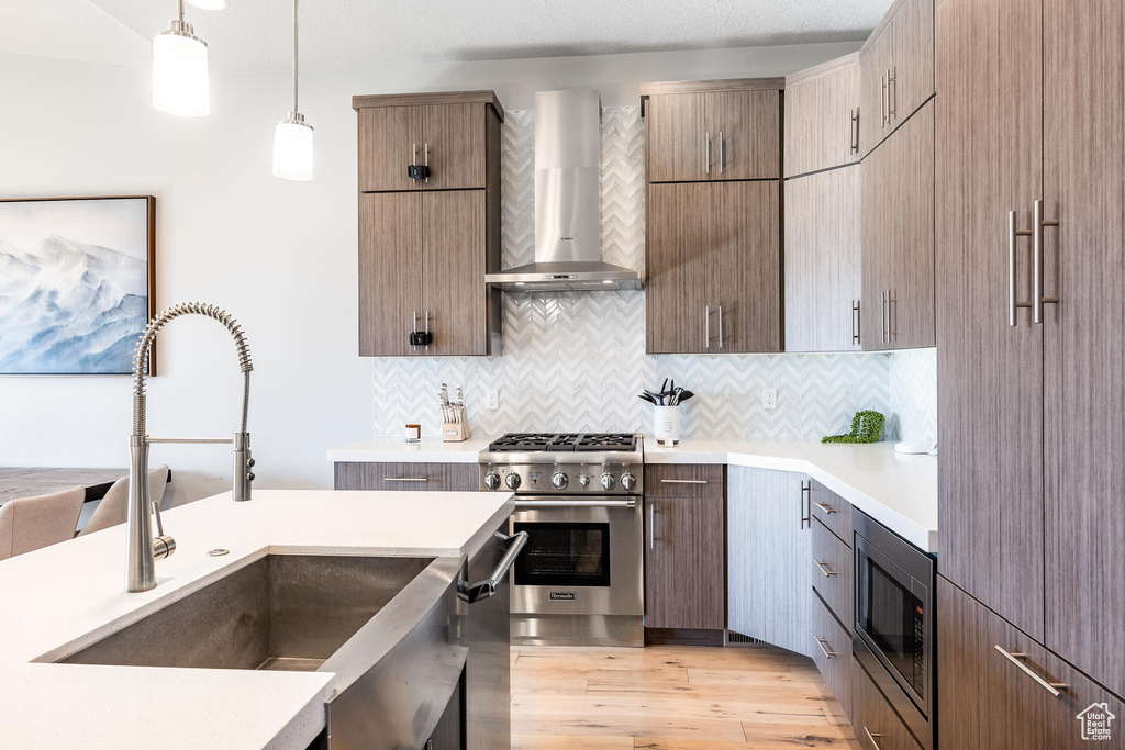 Kitchen featuring backsplash, light wood-type flooring, hanging light fixtures, stainless steel appliances, and wall chimney exhaust hood
