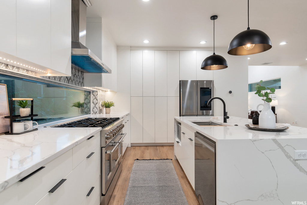 Kitchen featuring wall chimney exhaust hood, appliances with stainless steel finishes, white cabinets, decorative light fixtures, and backsplash