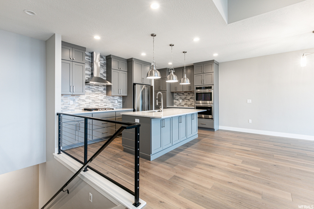 Kitchen with wall chimney range hood, appliances with stainless steel finishes, light hardwood / wood-style floors, tasteful backsplash, and an island with sink