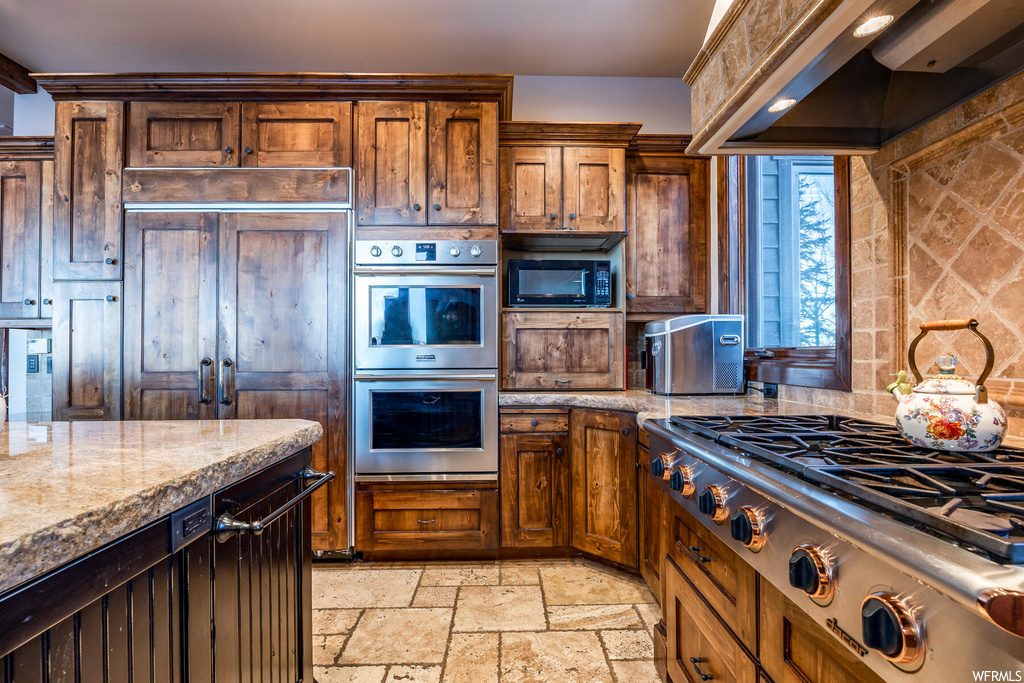 Kitchen with custom range hood, tasteful backsplash, light stone counters, built in appliances, and light tile floors