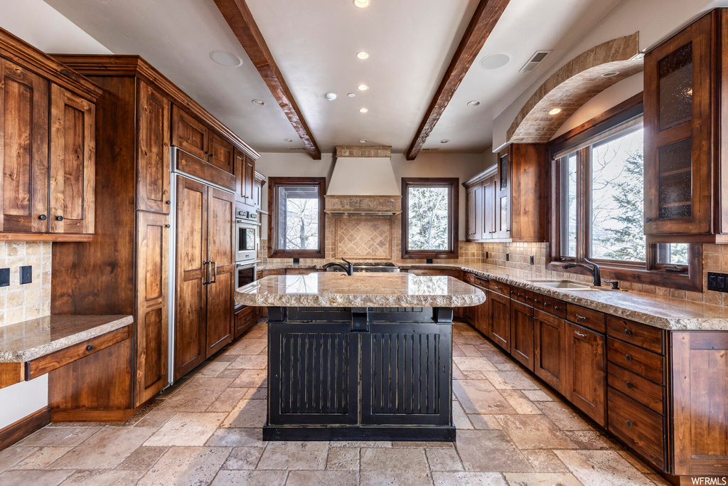 Kitchen with light tile floors, custom range hood, a kitchen island with sink, beamed ceiling, and backsplash