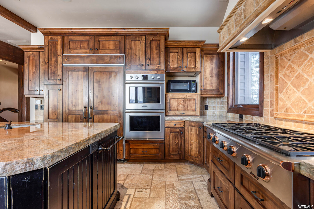 Kitchen with light stone counters, light tile flooring, built in appliances, custom exhaust hood, and backsplash