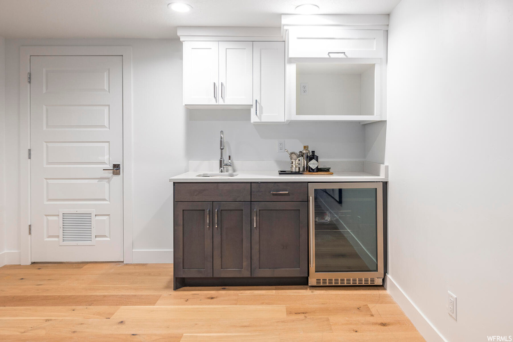 Bar featuring sink, dark brown cabinets, wine cooler, white cabinets, and light hardwood / wood-style flooring