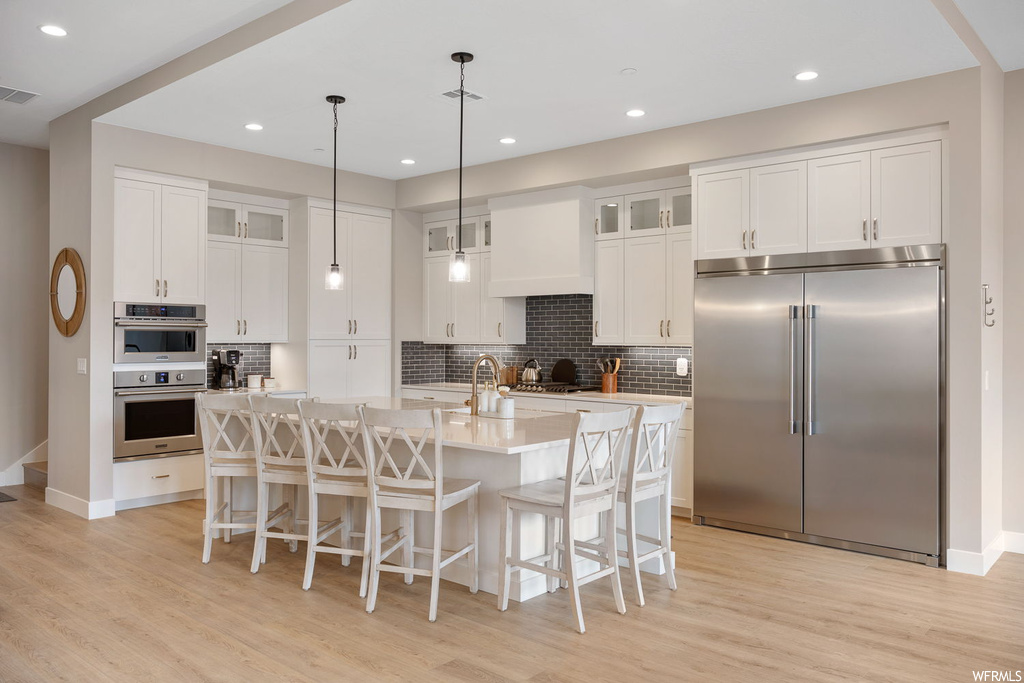 Kitchen featuring an island with sink, light hardwood / wood-style floors, appliances with stainless steel finishes, white cabinets, and decorative light fixtures