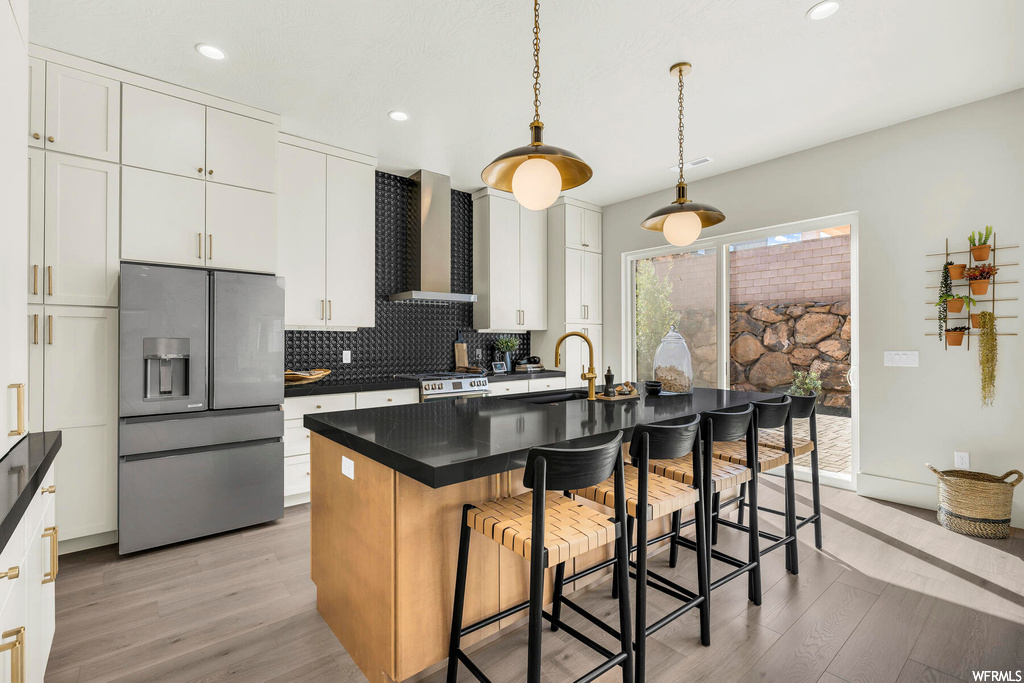 Kitchen featuring appliances with stainless steel finishes, wall chimney range hood, a kitchen island with sink, white cabinets, and light hardwood / wood-style flooring