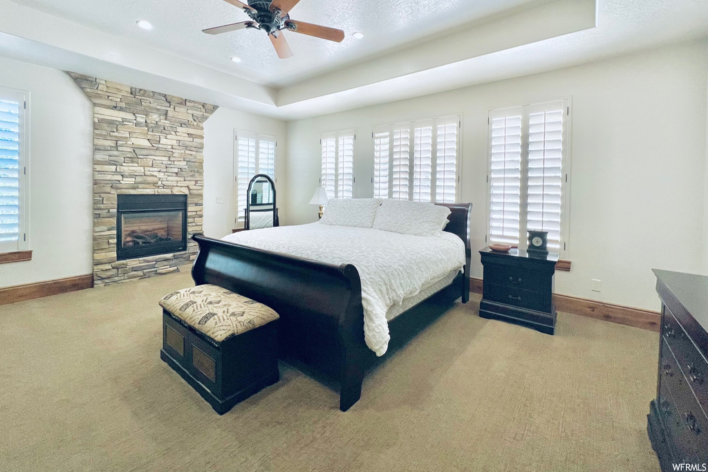 Bedroom featuring ceiling fan, a tray ceiling, a stone fireplace, and light carpet