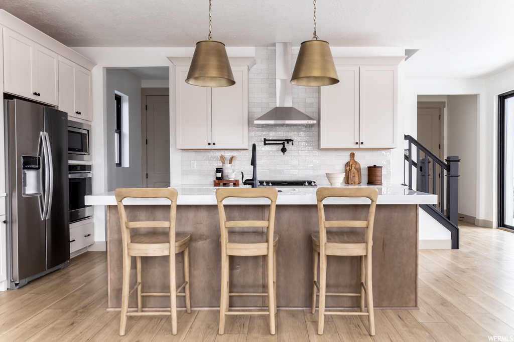 Kitchen with wall chimney range hood, pendant lighting, a breakfast bar area, stainless steel appliances, and a center island with sink