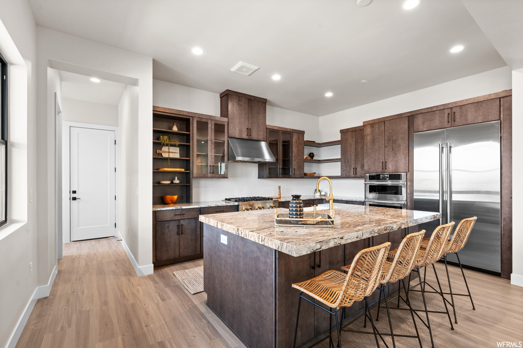 Kitchen with light hardwood / wood-style floors, dark brown cabinetry, stainless steel appliances, a center island with sink, and a kitchen breakfast bar