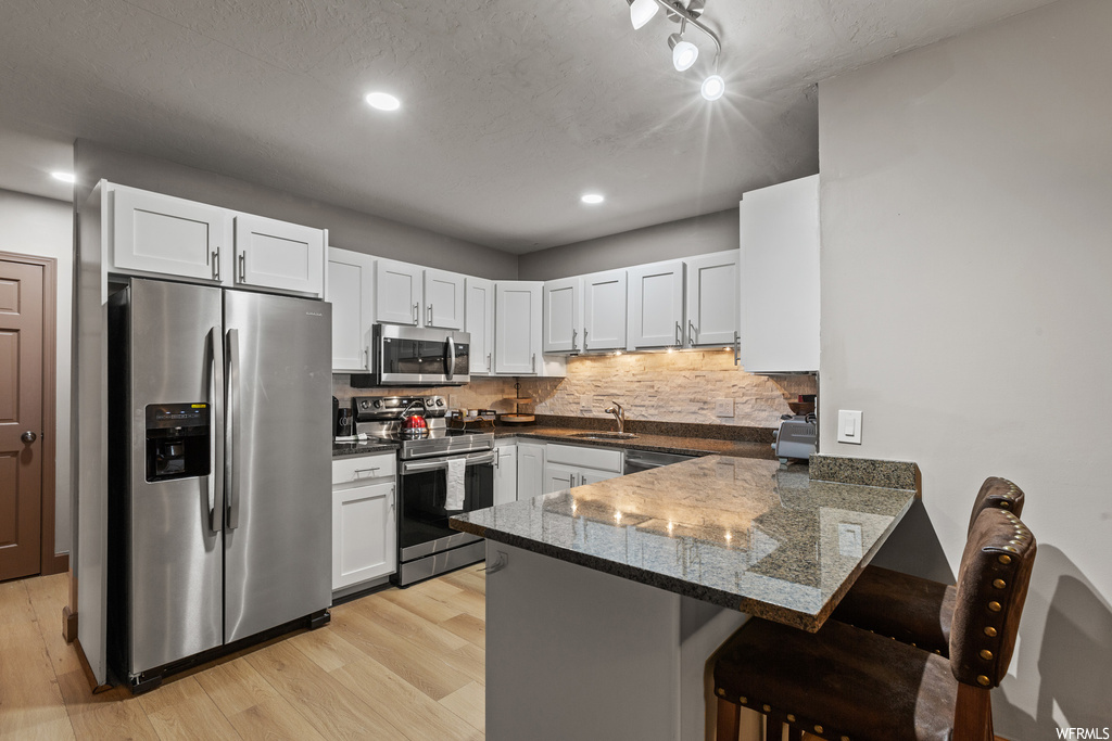 Kitchen with appliances with stainless steel finishes, backsplash, light hardwood / wood-style flooring, white cabinetry, and kitchen peninsula