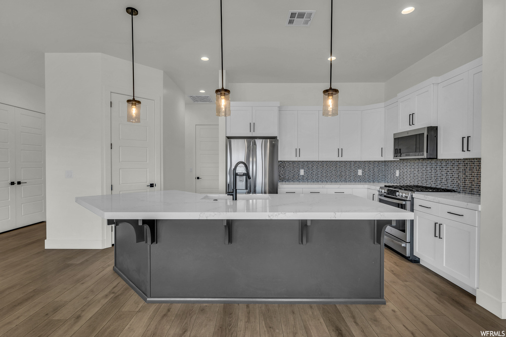 Kitchen with hanging light fixtures, a kitchen island with sink, stainless steel appliances, white cabinets, and wood-type flooring