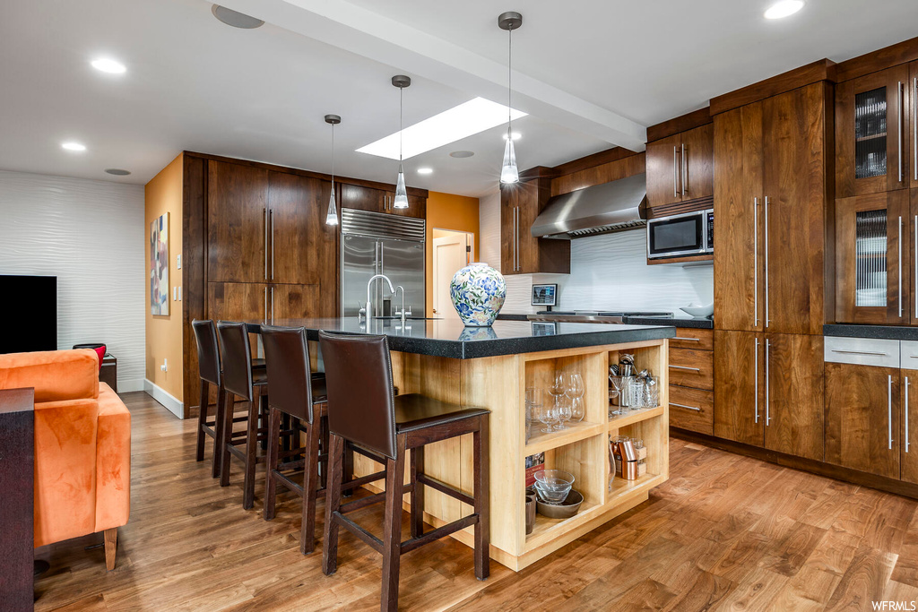 Kitchen featuring a kitchen island with sink, built in appliances, pendant lighting, light hardwood / wood-style floors, and wall chimney exhaust hood