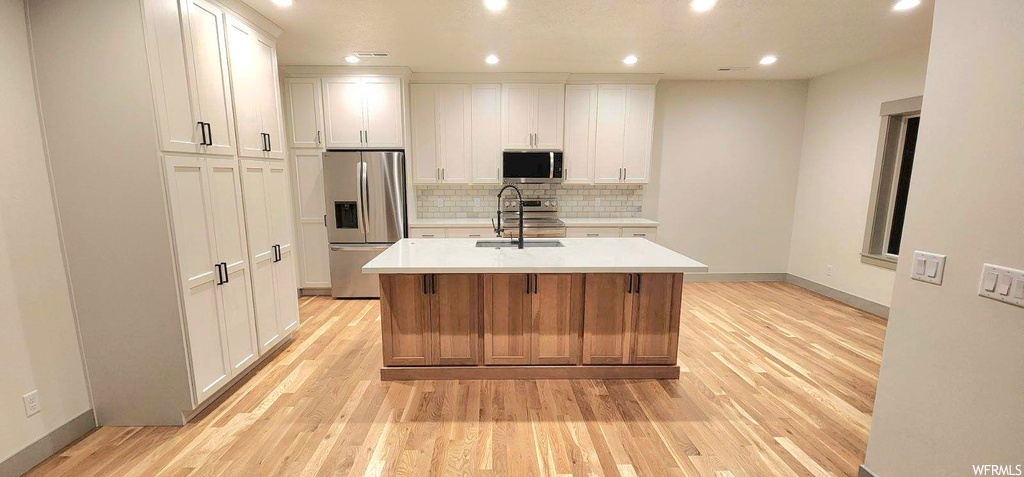 Kitchen with an island with sink, light wood-type flooring, white cabinets, and appliances with stainless steel finishes