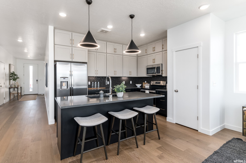 Kitchen featuring decorative light fixtures, light hardwood / wood-style flooring, stainless steel appliances, backsplash, and a kitchen island with sink