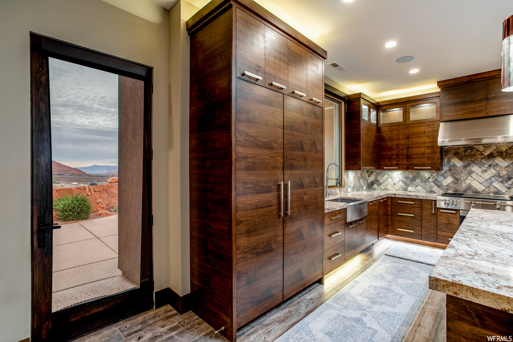 Kitchen featuring wall chimney exhaust hood, sink, dark brown cabinets, light stone countertops, and backsplash