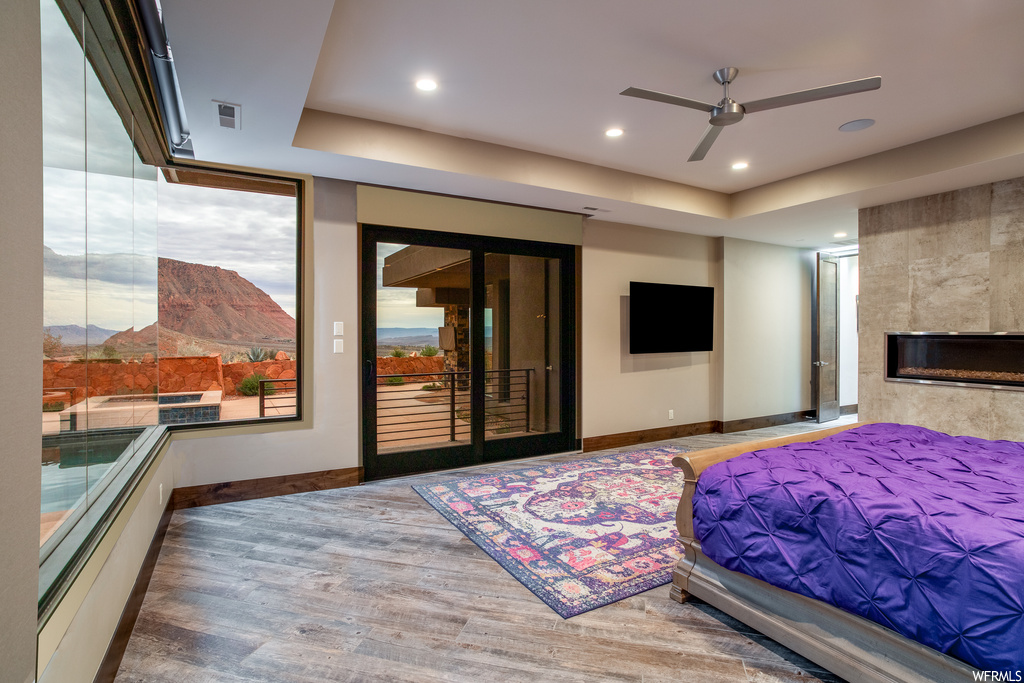 Bedroom with access to outside, a tile fireplace, light hardwood / wood-style floors, ceiling fan, and a raised ceiling