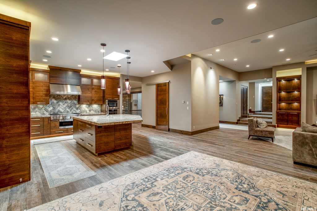 Kitchen with wall chimney exhaust hood, hanging light fixtures, a center island, backsplash, and light wood-type flooring