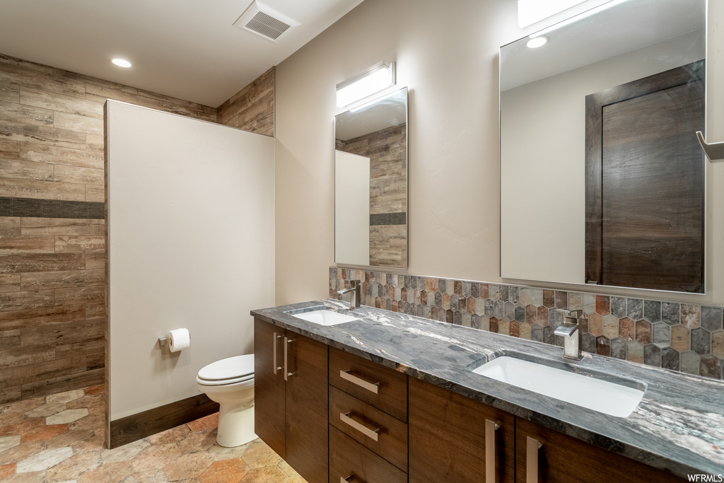 Bathroom with toilet, dual bowl vanity, tile flooring, and backsplash