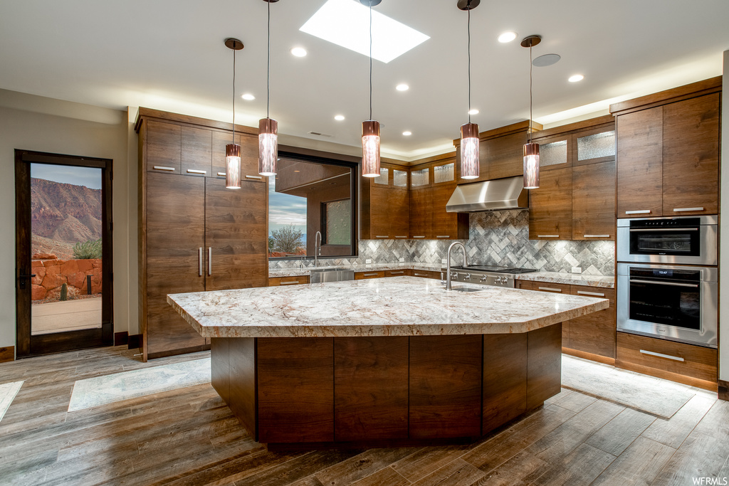 Kitchen featuring wall chimney range hood, a kitchen island with sink, hanging light fixtures, appliances with stainless steel finishes, and backsplash