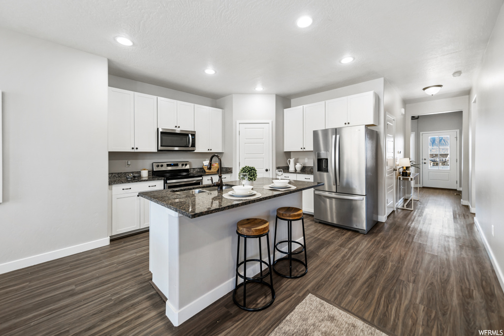 Kitchen featuring stainless steel appliances, white cabinetry, an island with sink, and dark wood-type flooring