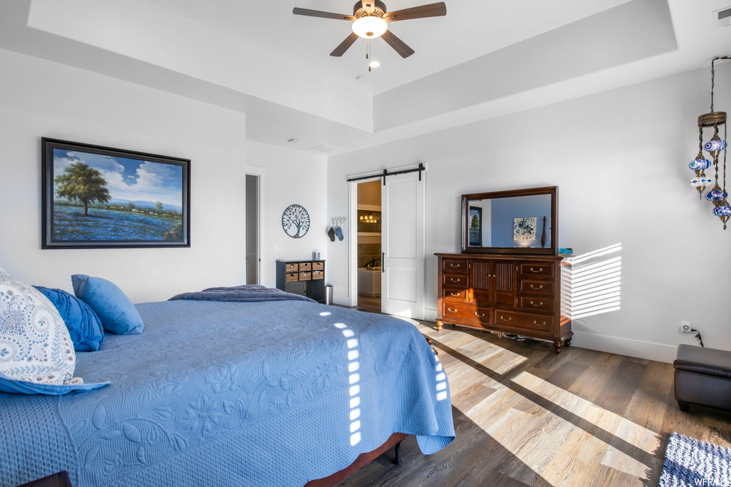 Bedroom with a barn door, a raised ceiling, dark hardwood / wood-style floors, and ceiling fan