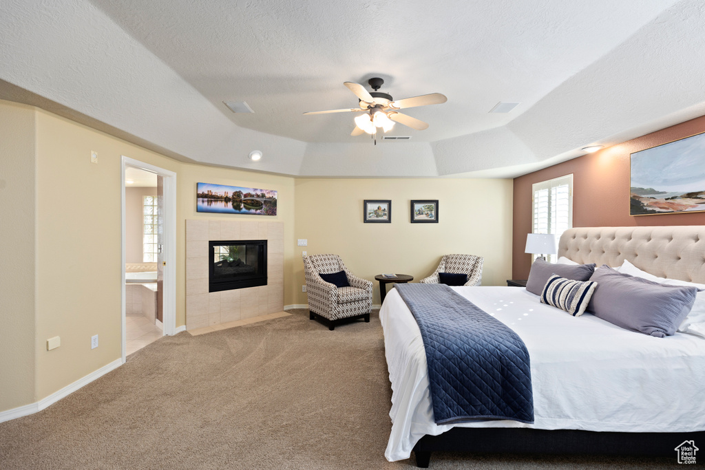Carpeted bedroom with connected bathroom, a tiled fireplace, a tray ceiling, and ceiling fan