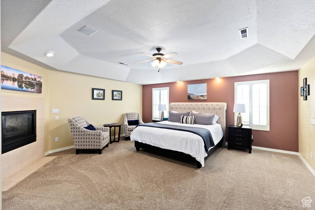 Carpeted bedroom with multiple windows, a tiled fireplace, a tray ceiling, and ceiling fan