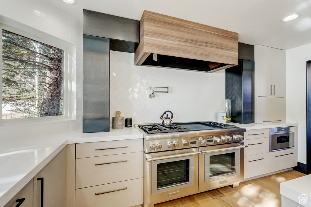 Kitchen with custom exhaust hood, stainless steel appliances, light hardwood / wood-style floors, and backsplash