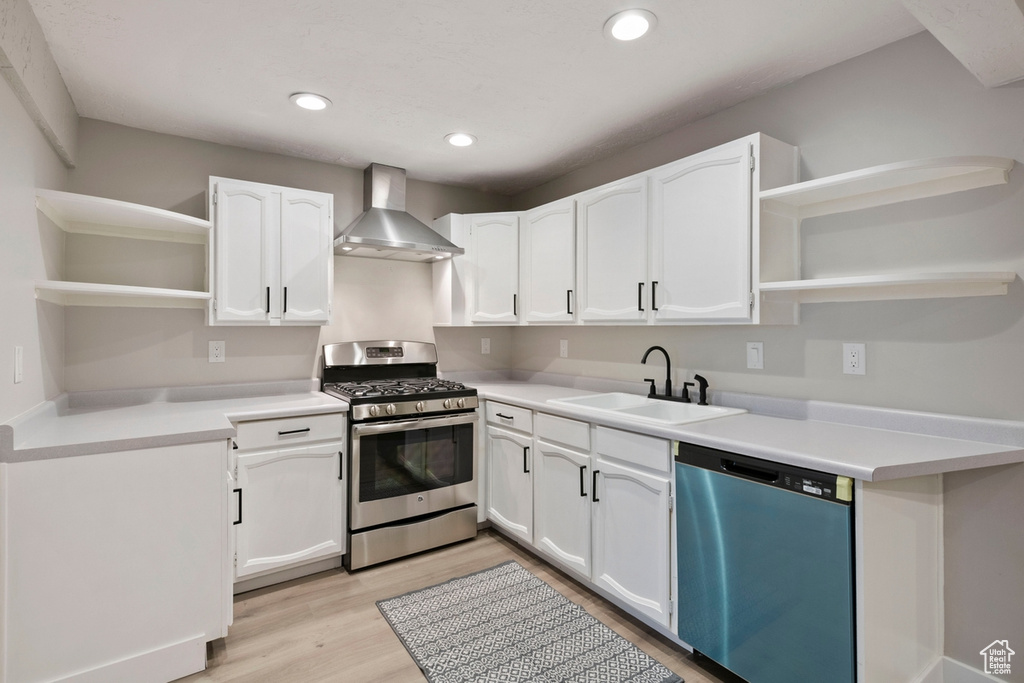 Kitchen with appliances with stainless steel finishes, white cabinets, sink, light hardwood / wood-style floors, and wall chimney range hood