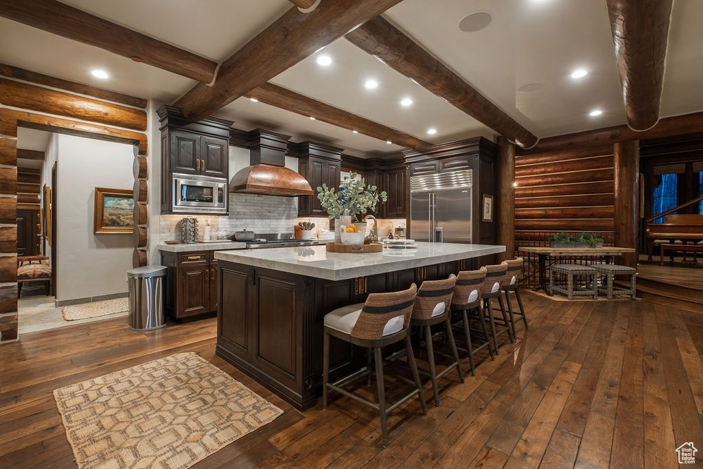 Kitchen with tasteful backsplash, dark wood-type flooring, a kitchen island with sink, custom exhaust hood, and built in appliances