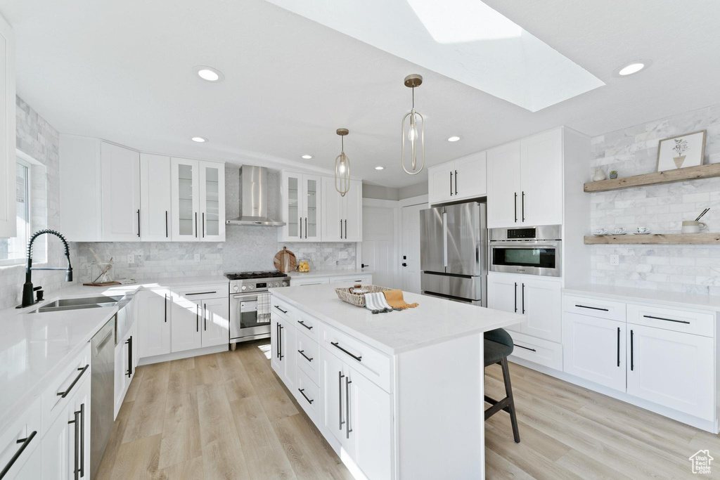 Kitchen featuring a skylight, wall chimney range hood, stainless steel appliances, and light wood-type flooring