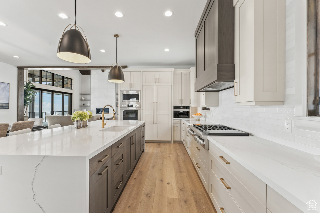 Kitchen featuring decorative light fixtures, premium range hood, an island with sink, sink, and light wood-type flooring