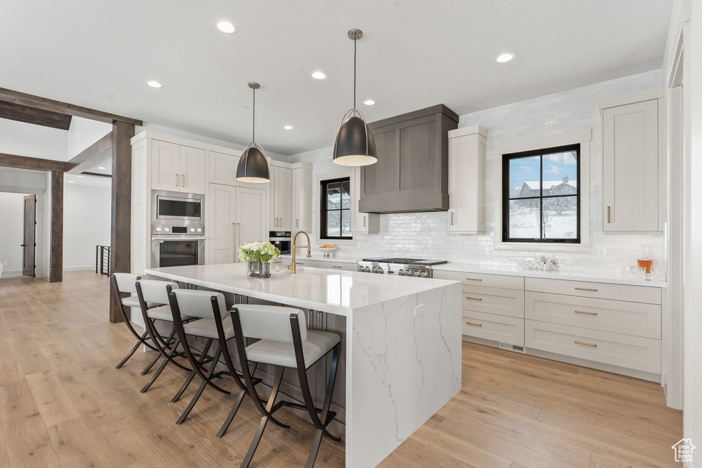 Kitchen featuring decorative light fixtures, light hardwood / wood-style flooring, premium range hood, an island with sink, and stainless steel microwave