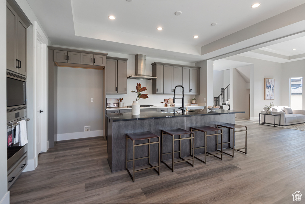 Kitchen featuring dark hardwood / wood-style flooring, a tray ceiling, a center island with sink, wall chimney range hood, and stainless steel appliances