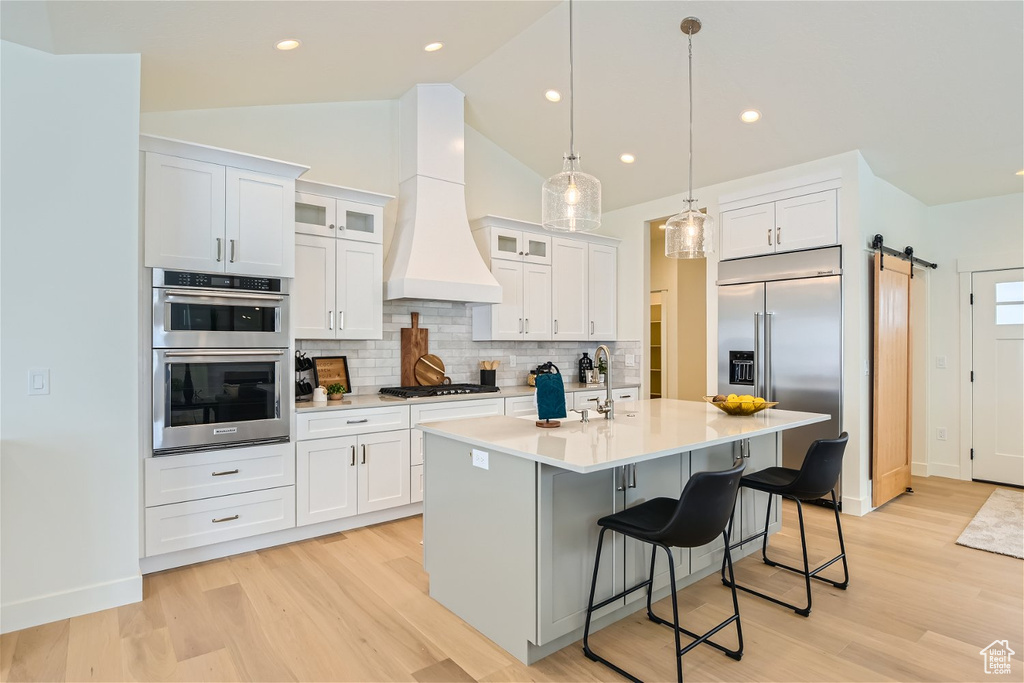 Kitchen featuring premium range hood, light wood-type flooring, appliances with stainless steel finishes, a barn door, and an island with sink
