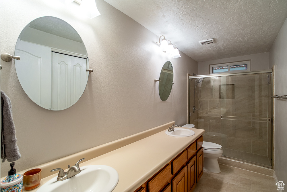 Bathroom featuring a textured ceiling, toilet, double vanity, a shower with shower door, and tile flooring