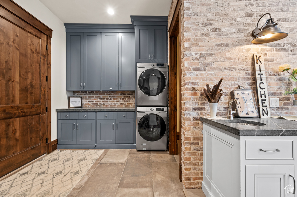 Clothes washing area with light tile floors, cabinets, brick wall, and stacked washer and dryer