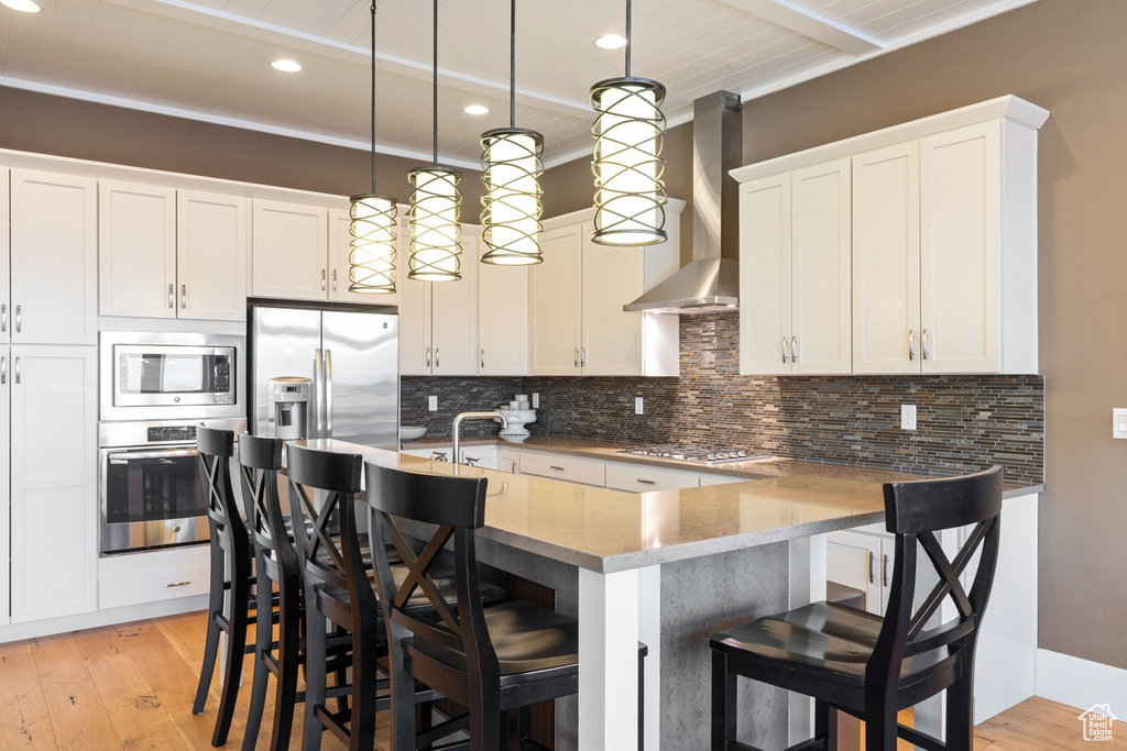 Kitchen with appliances with stainless steel finishes, wall chimney range hood, light hardwood / wood-style flooring, and decorative light fixtures