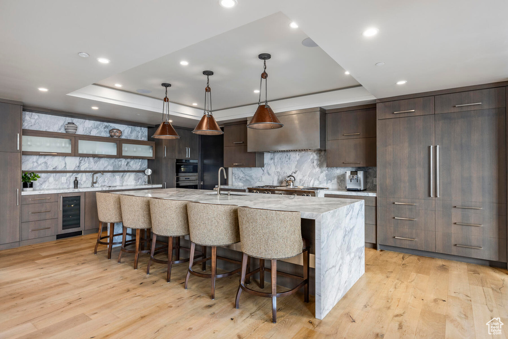Kitchen with light hardwood / wood-style floors, tasteful backsplash, wall chimney exhaust hood, and a kitchen island with sink