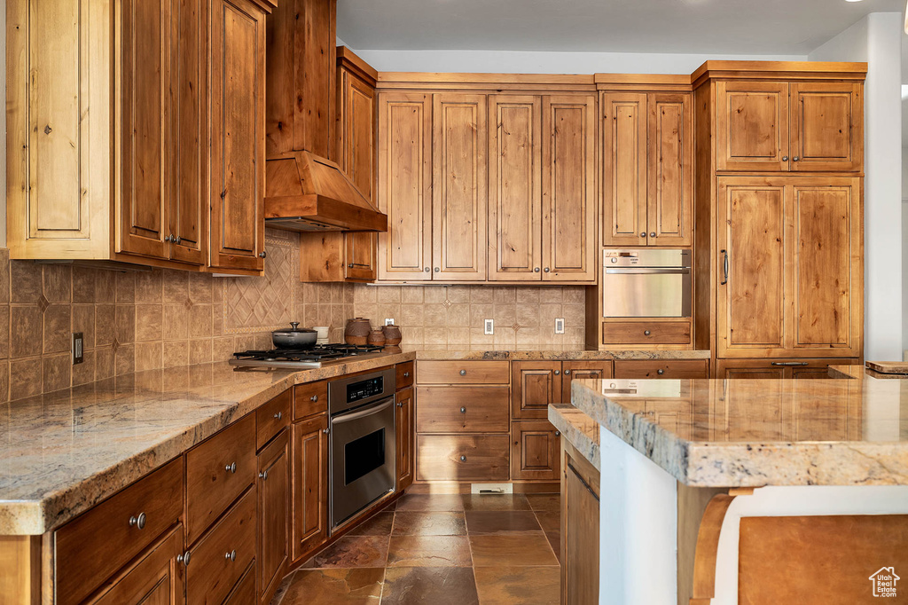 Kitchen featuring appliances with stainless steel finishes, premium range hood, light stone counters, dark tile floors, and tasteful backsplash
