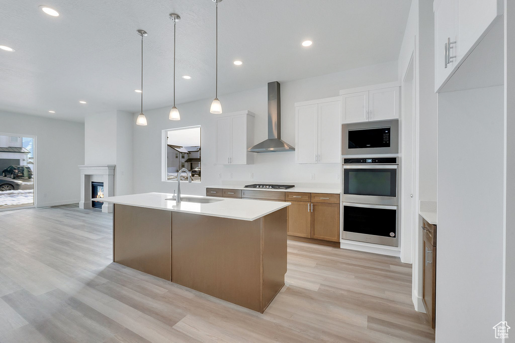 Kitchen featuring white cabinetry, light wood-type flooring, a kitchen island with sink, sink, and wall chimney range hood