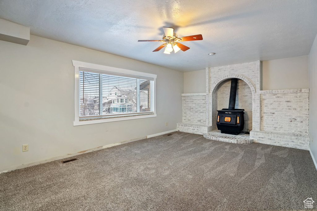 Unfurnished living room featuring a wood stove, a textured ceiling, brick wall, ceiling fan, and carpet floors