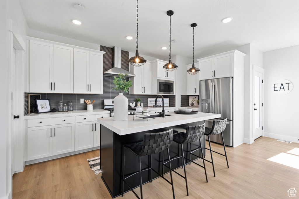 Kitchen featuring backsplash, light hardwood / wood-style flooring, a center island with sink, wall chimney range hood, and stainless steel appliances