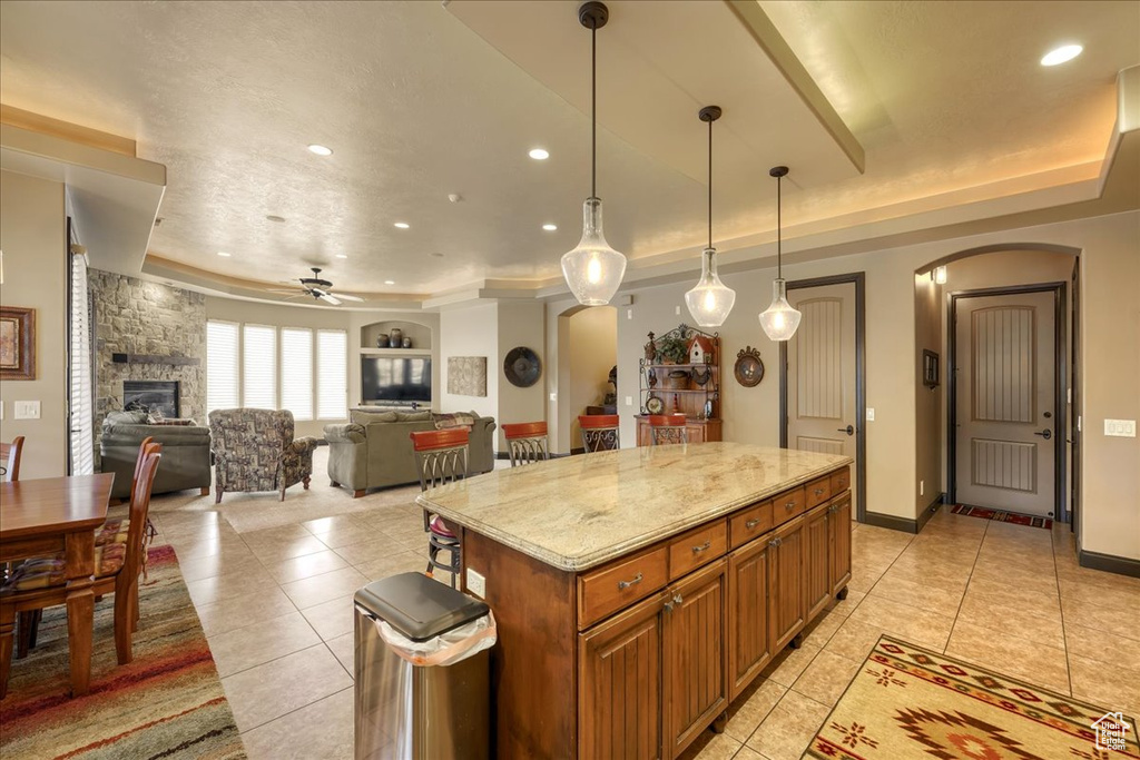 Kitchen featuring a center island, a stone fireplace, light stone counters, ceiling fan, and a tray ceiling