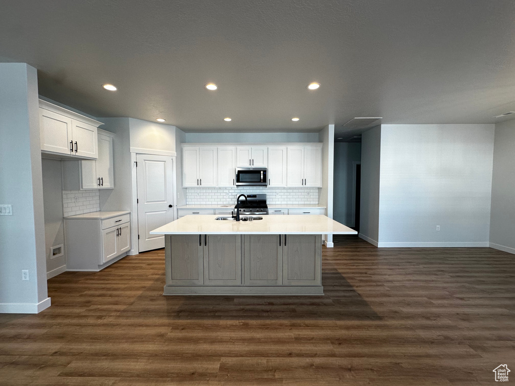 Kitchen with white cabinets, stainless steel appliances, an island with sink, backsplash, and dark wood-type flooring