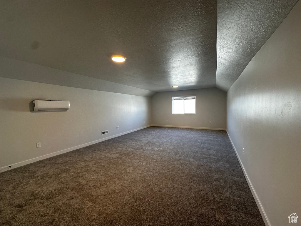 Additional living space with a wall mounted AC, vaulted ceiling, a textured ceiling, and dark colored carpet