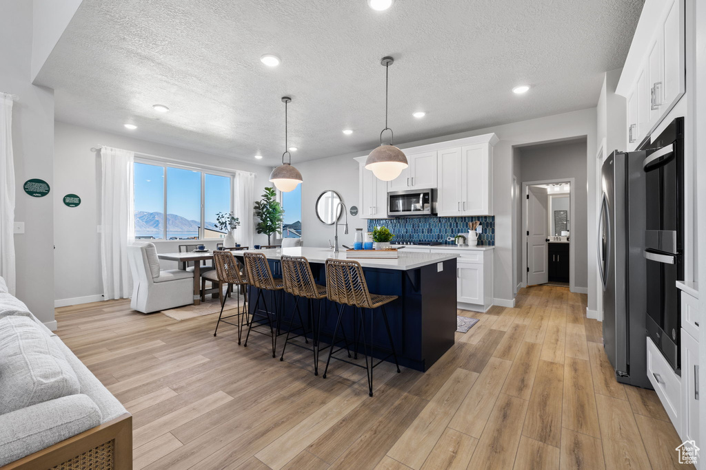 Kitchen featuring decorative light fixtures, white cabinets, light hardwood / wood-style flooring, stainless steel appliances, and a kitchen island with sink