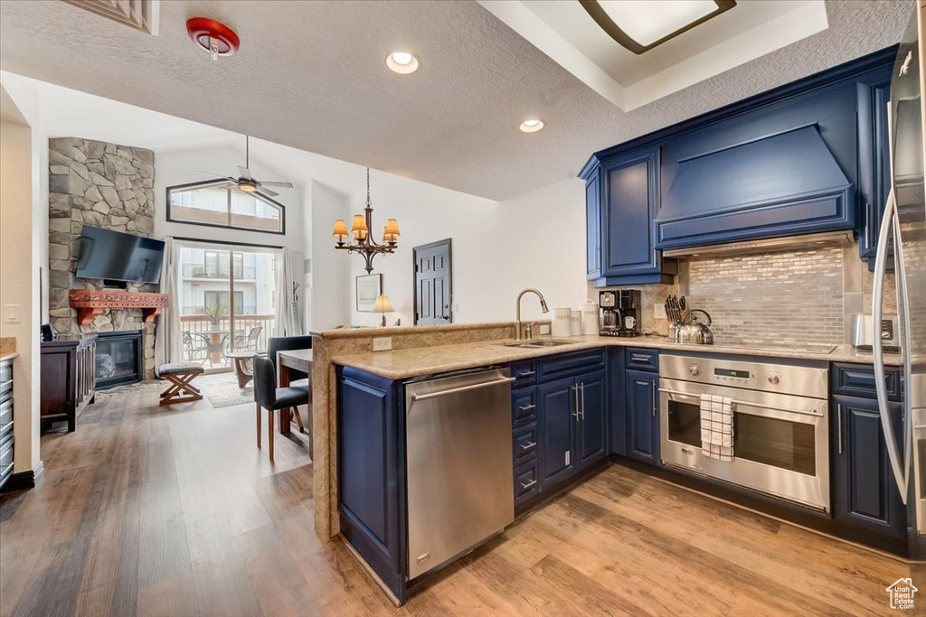 Kitchen with premium range hood, ceiling fan with notable chandelier, appliances with stainless steel finishes, light hardwood / wood-style flooring, and a fireplace